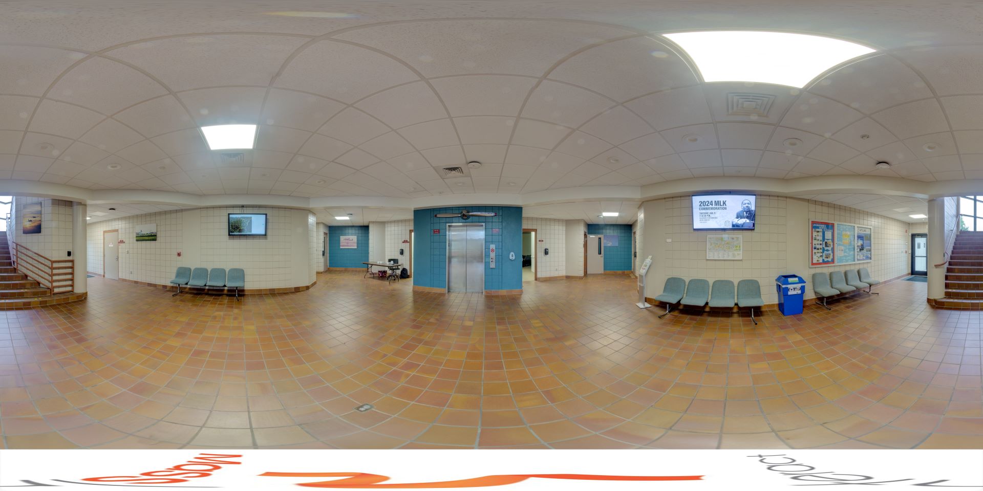 Panoramic view of a spacious lobby with tiled flooring, featuring rows of blue chairs, multiple doors, and a curved ceiling, flanked by staircases on both sides and decorated with informational posters